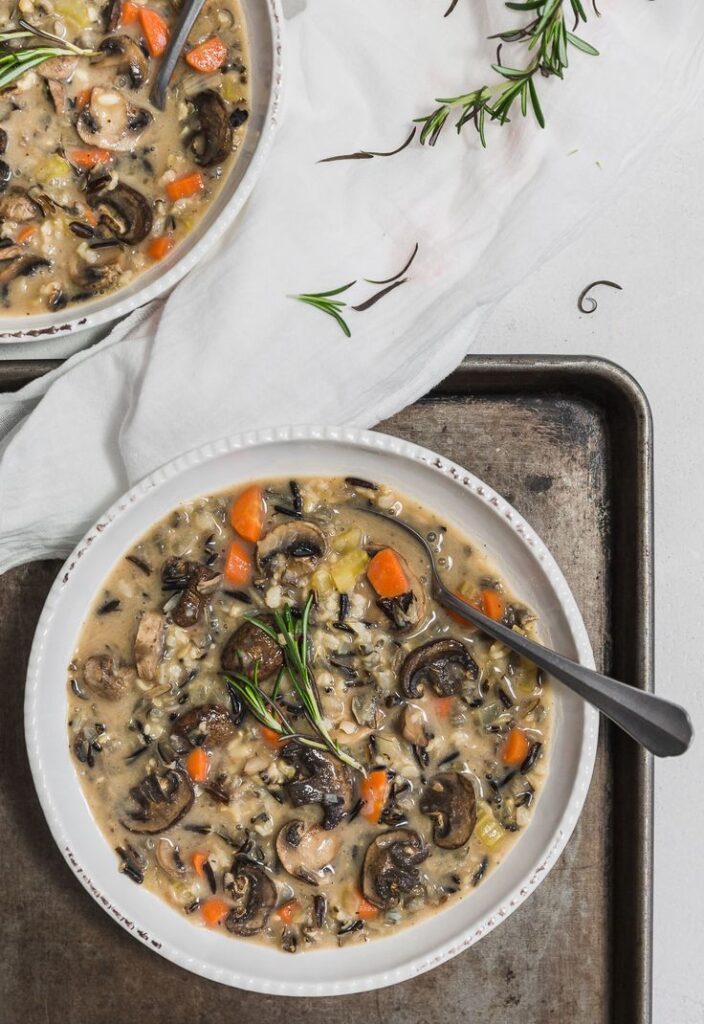Creamy wild rice soup with mushrooms and carrots in a bowl, garnished with fresh rosemary on a rustic tray.
