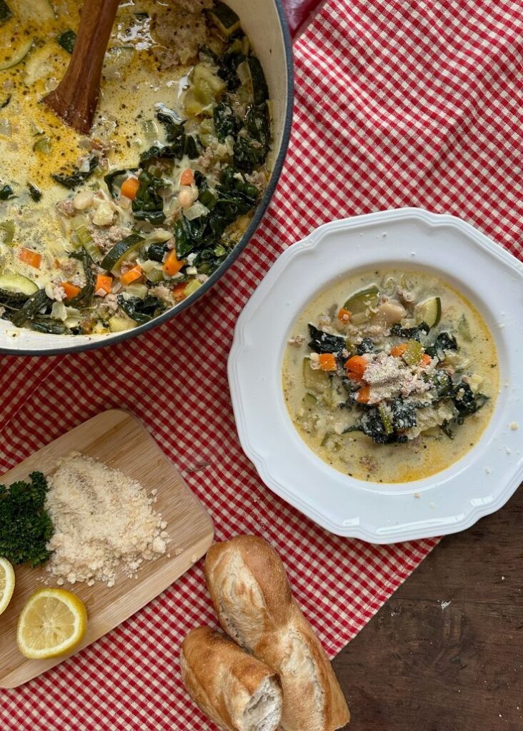 Creamy vegetable soup in a pot and bowl with bread, cheese, parsley, and lemon on a red checkered cloth.