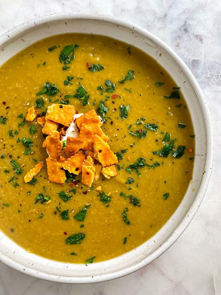 Creamy yellow lentil soup garnished with fresh herbs and crushed crackers in a white bowl on a marble surface.