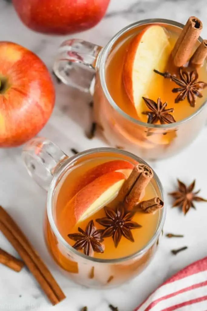Two mugs of spiced apple cider with cinnamon sticks, star anise, and apple slices on a marble countertop.