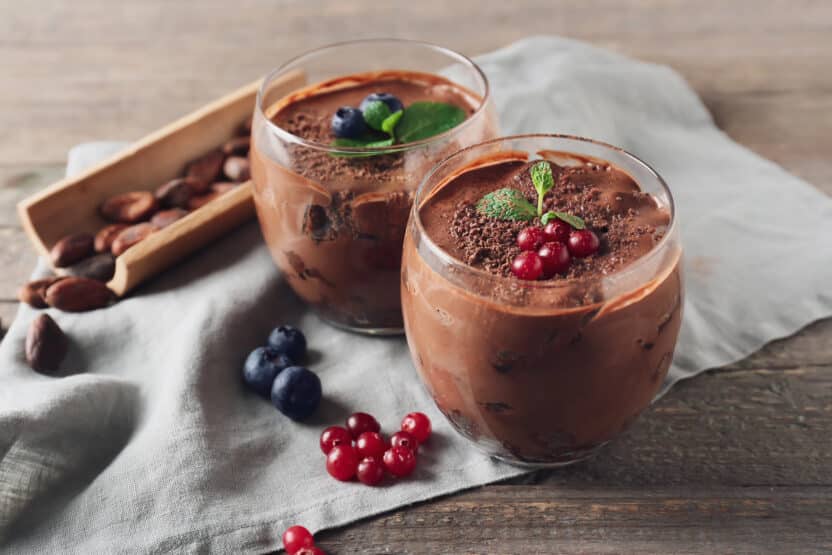 Two glasses of rich chocolate mousse topped with fresh berries and mint leaves on a wooden table.