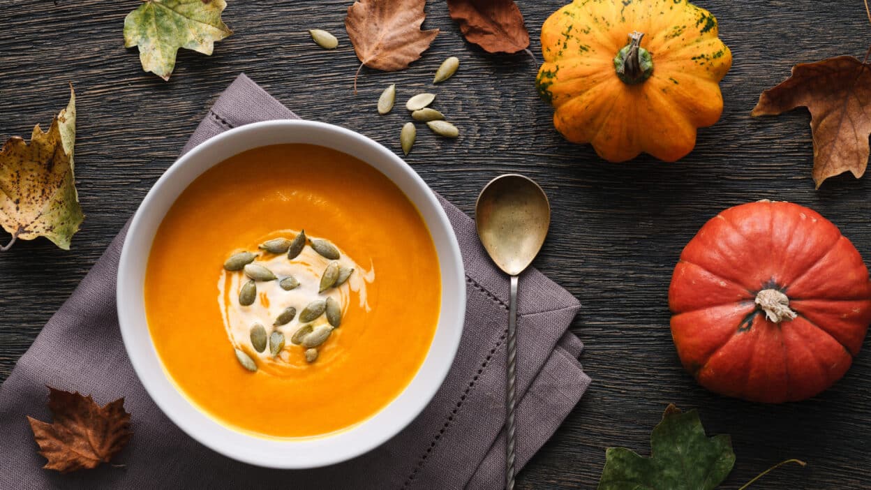 Creamy pumpkin soup garnished with seeds, surrounded by fall leaves and pumpkins on a rustic wooden table.