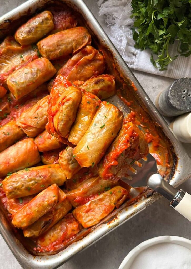 Stuffed cabbage rolls in tomato sauce on a tray with fresh herbs and a serving spatula.