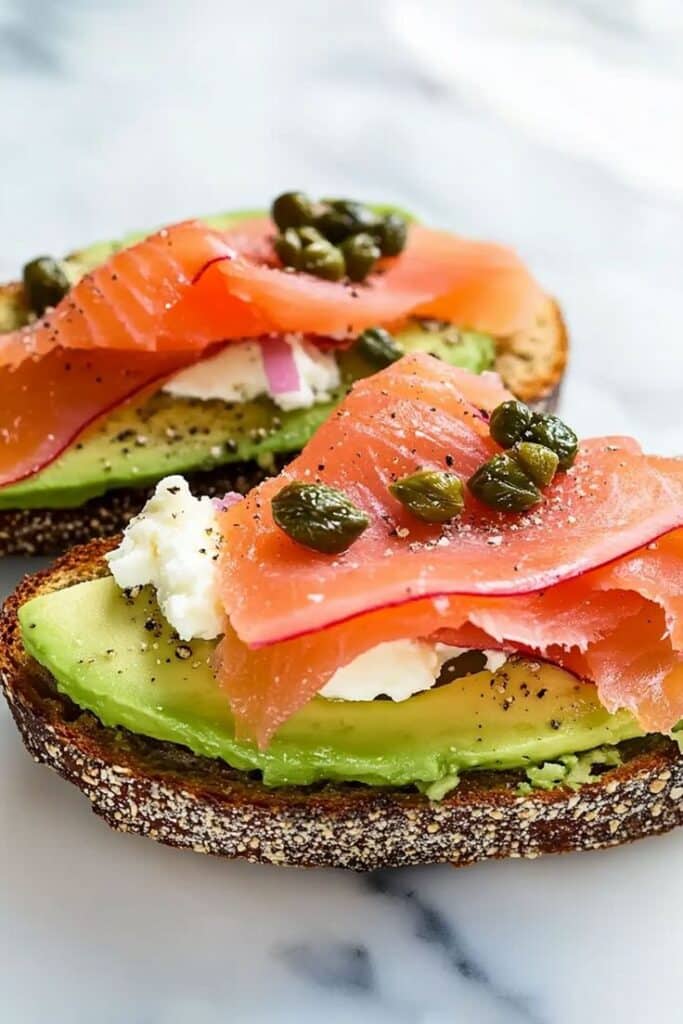 Smoked salmon, avocado, cream cheese, and capers on toast, garnished with black pepper, served on a marble surface.