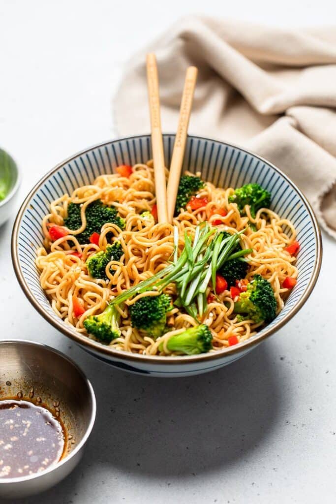 Bowl of noodles with broccoli and peppers, topped with scallions, with chopsticks on a light background.