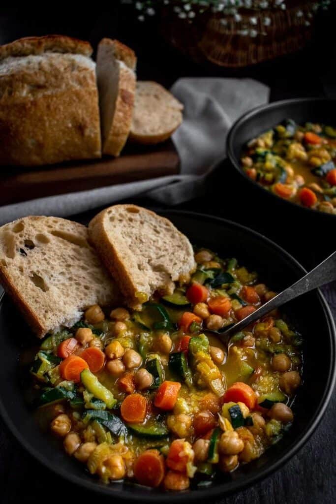 Rustic vegetable soup with chickpeas and slices of bread on a dark table setting.