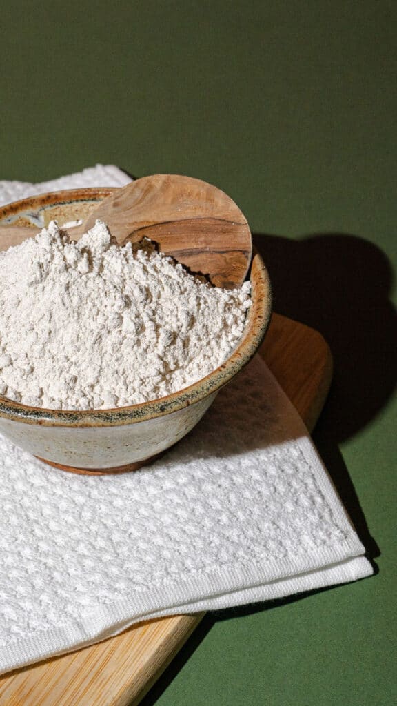 Gluten-Free Flour Alternative_ Fonio - Master the Art of Homemade Pizza with Fun, Simple Steps | Ceramic bowl with white powder and wooden scoop on white cloth, set on green surface.