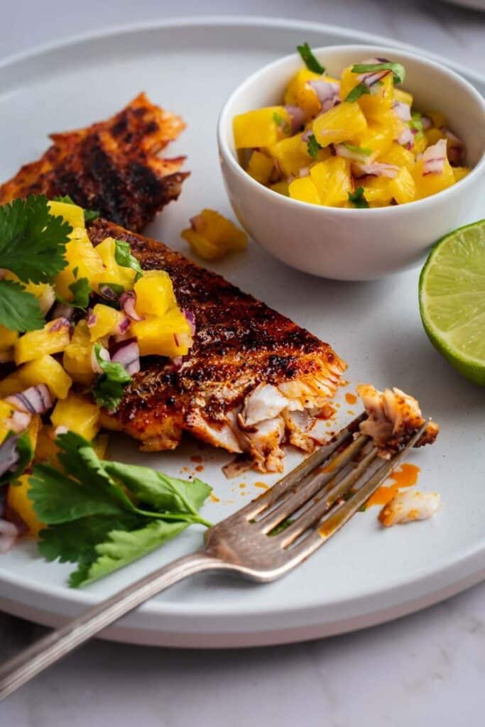 Grilled fish topped with mango salsa and cilantro, served with lime on a white plate.