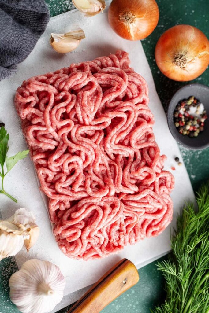 Fresh ground meat on a cutting board surrounded by onions, garlic, herbs, and peppercorns for cooking.