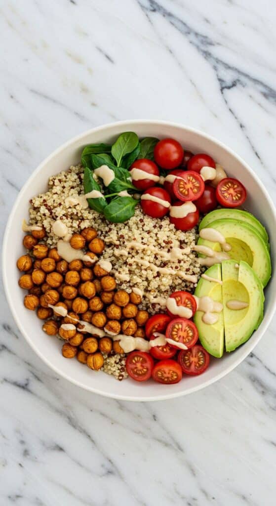 Healthy vegan bowl with quinoa, chickpeas, spinach, cherry tomatoes, avocado, and tahini drizzle on marble background.