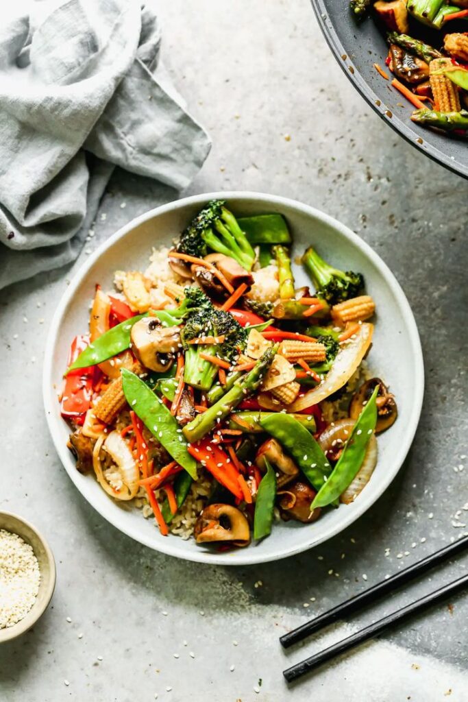 Colorful vegetable stir-fry with broccoli, peppers, and snow peas on rice in a white bowl, garnished with sesame seeds.