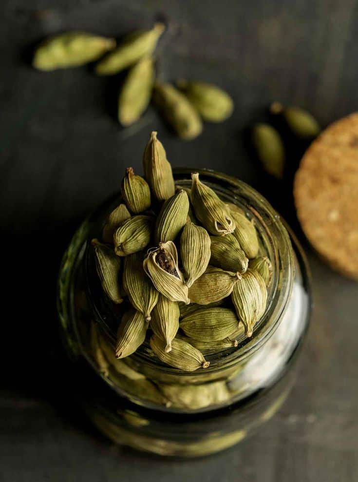 Jar filled with cardamom pods, with some scattered on a dark surface. Perfect for spice and cooking themes.