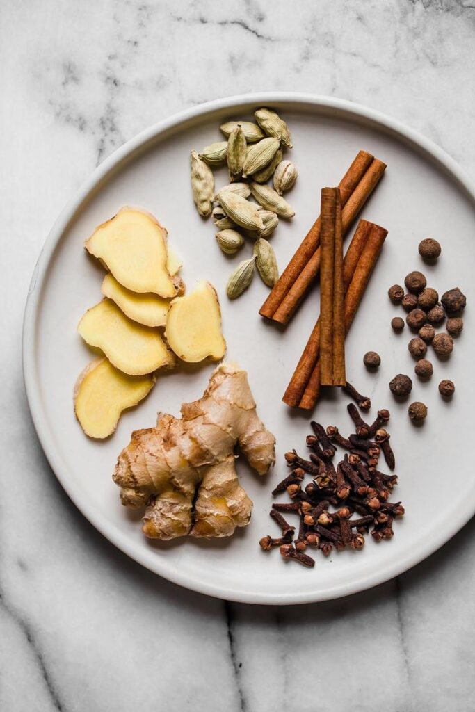 Assorted spices on a white plate: ginger, cardamom, cinnamon, allspice, and cloves on a marble background.