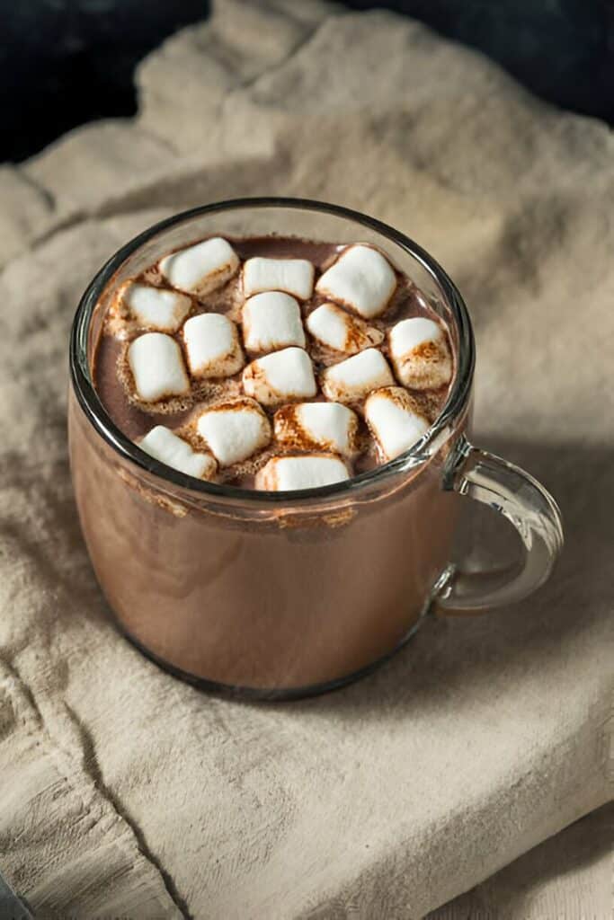 Hot chocolate with marshmallows in a glass mug on a beige cloth, cozy and delicious winter treat.