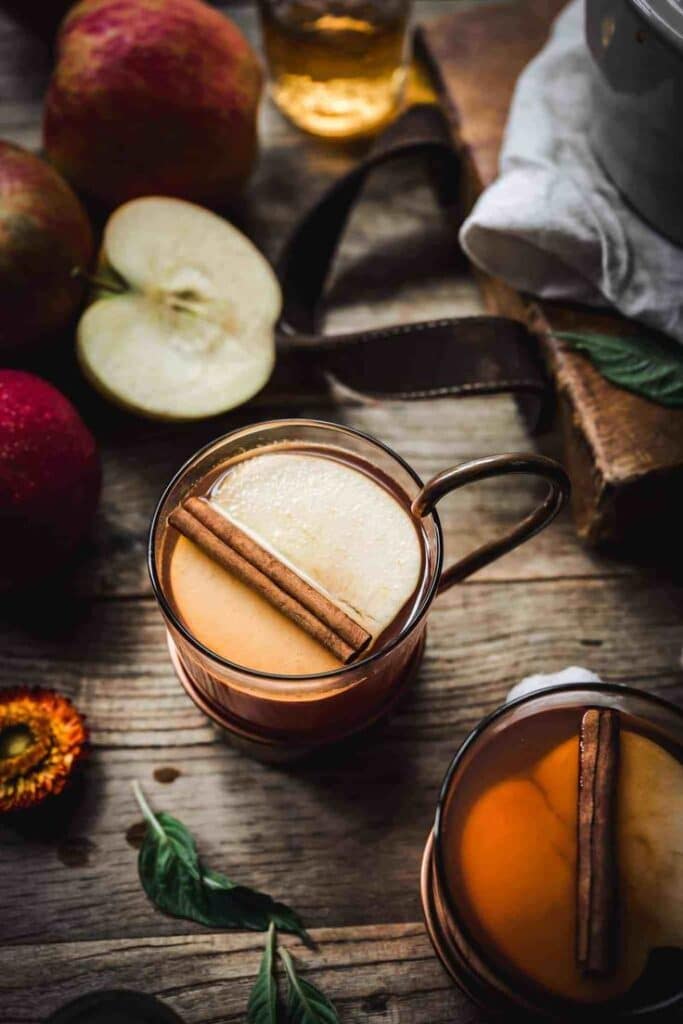 Warm apple cider with cinnamon sticks in glass mugs on wooden table, surrounded by fresh apples and greenery.
