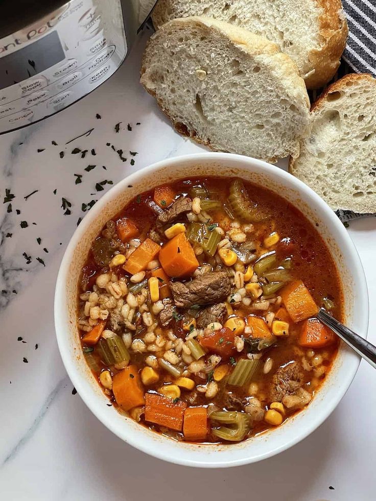 Hearty beef and barley soup with vegetables served in a white bowl, accompanied by sliced bread and sprinkled herbs.