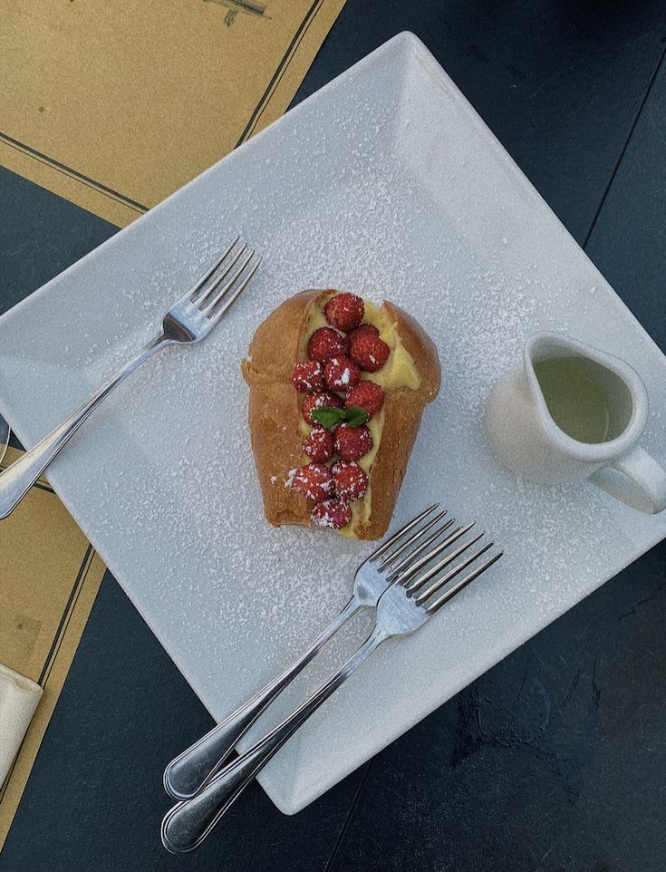 Dessert with strawberries and cream on a square plate, served with forks and a side of sauce on a dark table.