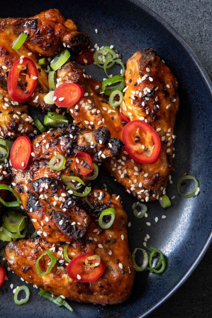 Spicy grilled chicken wings with sesame seeds and sliced red peppers on a black plate.