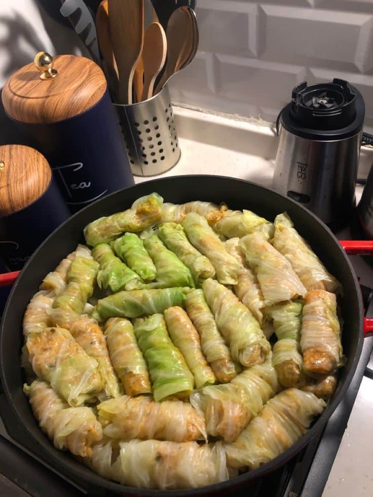 Stuffed cabbage rolls in a skillet on a countertop, surrounded by kitchen utensils and storage canisters.