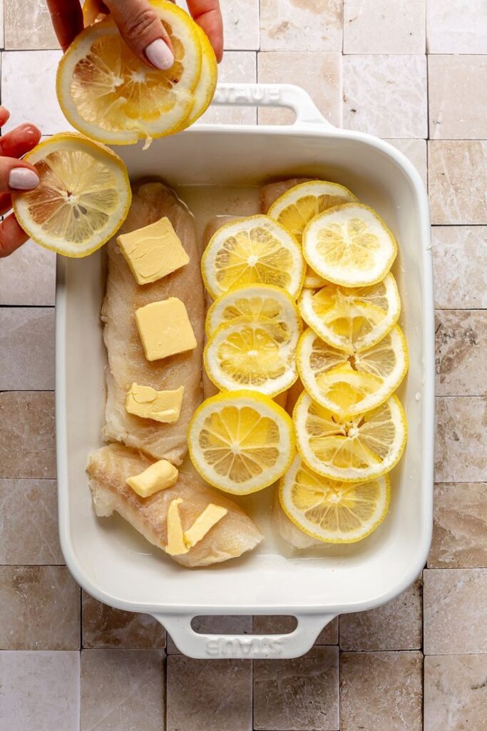 Baking dish with fish fillets topped with butter and lemon slices, ready for cooking on a tiled background.