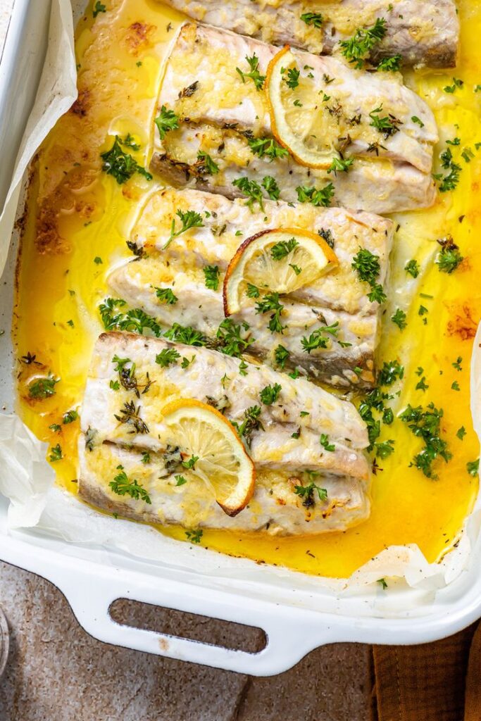 Baked fish fillets with lemon slices and fresh herbs in a white baking dish, perfect for a healthy dinner.