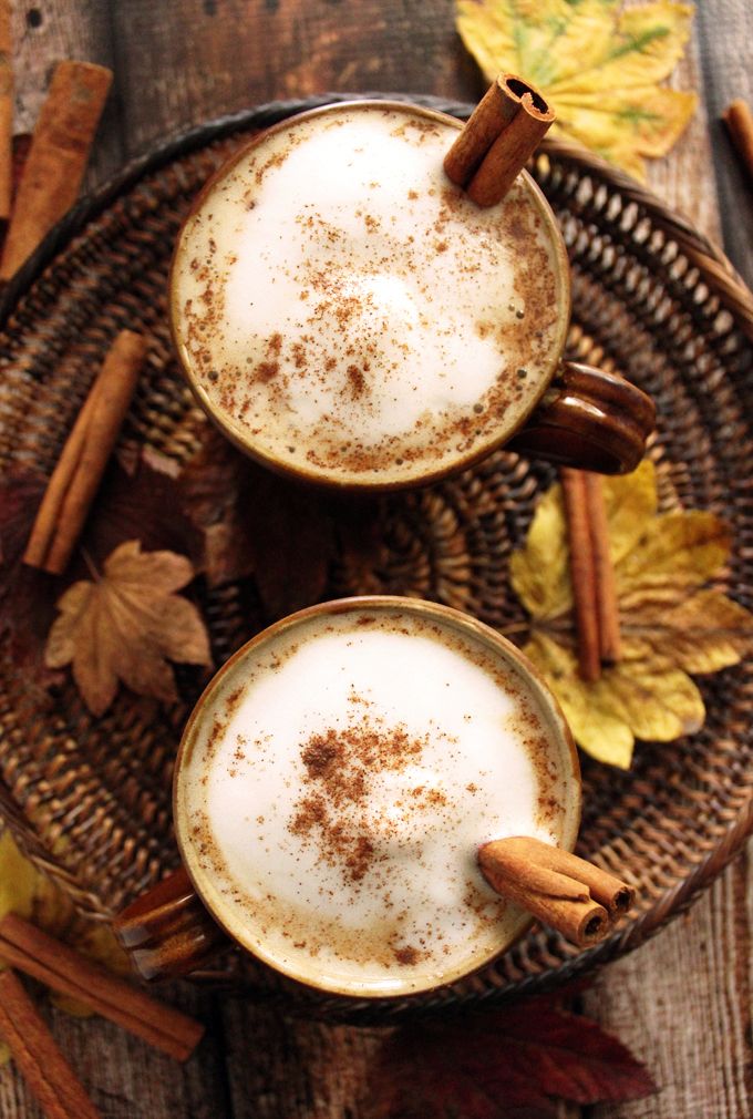 Two frothy chai lattes with cinnamon sticks on a rustic table, surrounded by autumn leaves and spices.