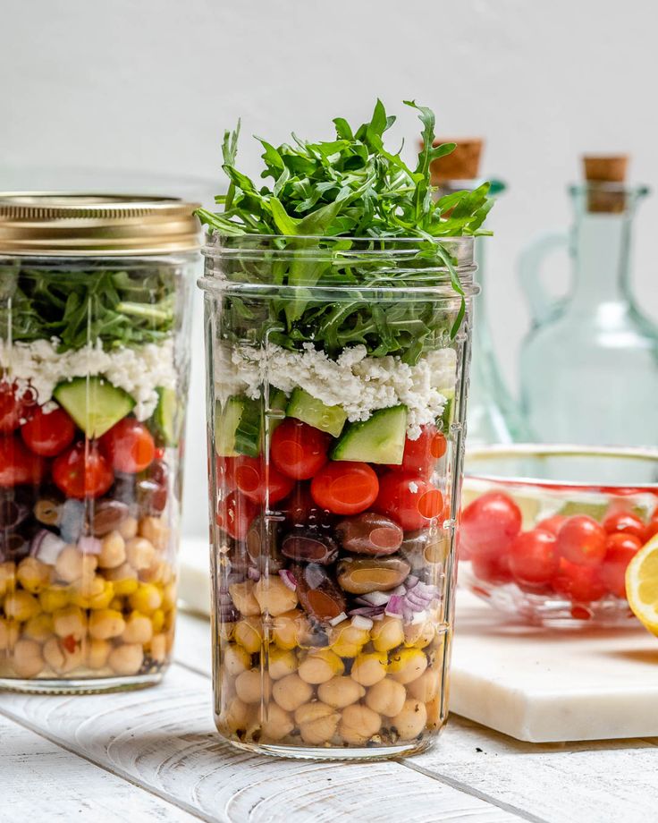 Layered Mediterranean salad in a jar with chickpeas, olives, tomatoes, cucumbers, feta, and arugula on a white surface.
