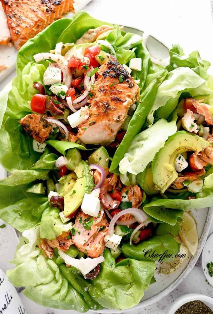 Grilled salmon lettuce wraps with avocado, onion, cheese, and tomato, garnished with herbs on a white plate.