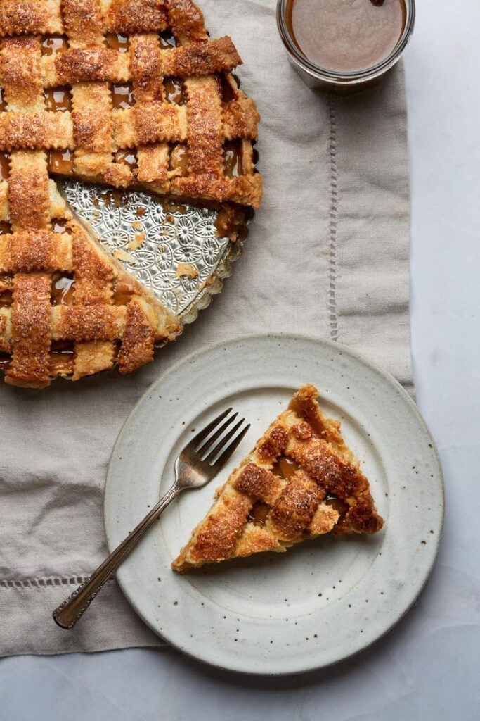 Slice of homemade lattice apple pie on a plate with fork, next to the full pie, on a linen cloth.