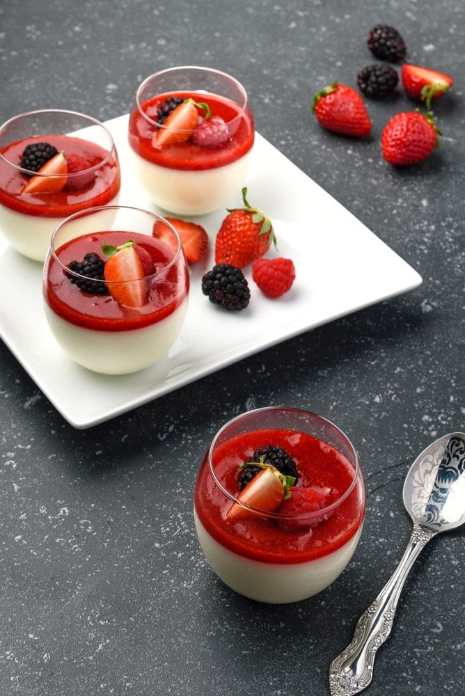 Creamy panna cotta in glass cups topped with fresh berries on a dark surface with a vintage spoon nearby.