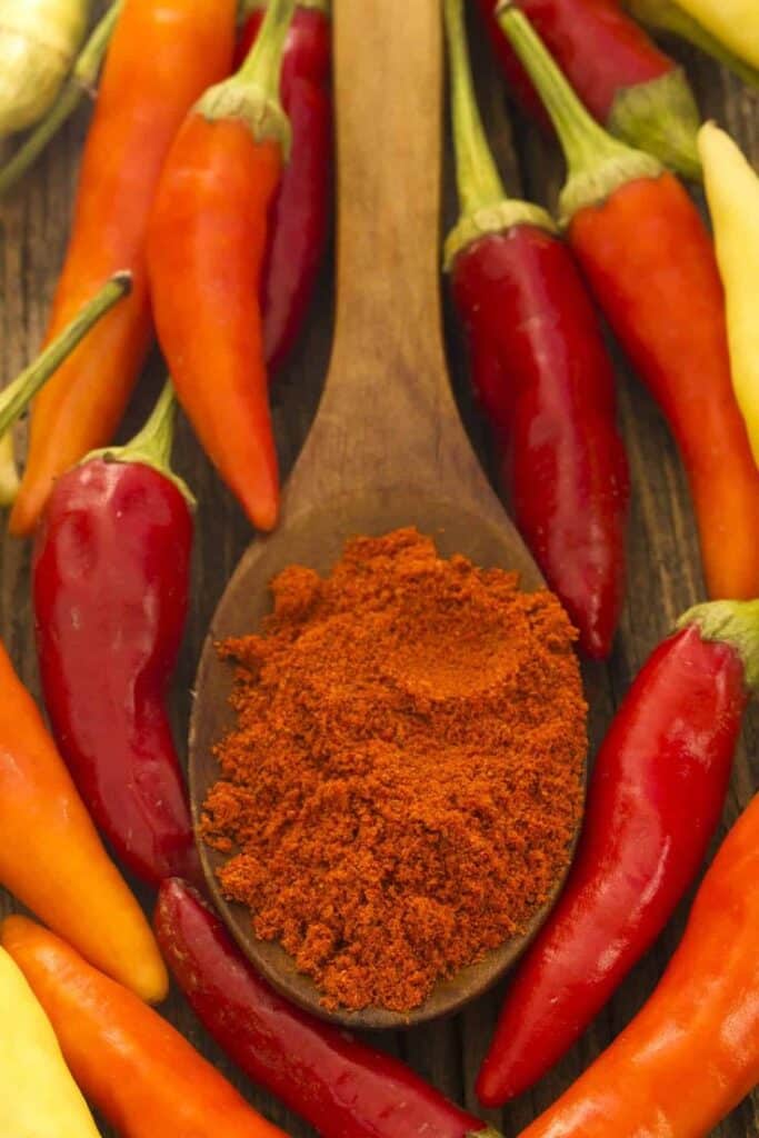 Assorted red and orange chili peppers with ground paprika on a wooden spoon, showcasing spicy flavors.