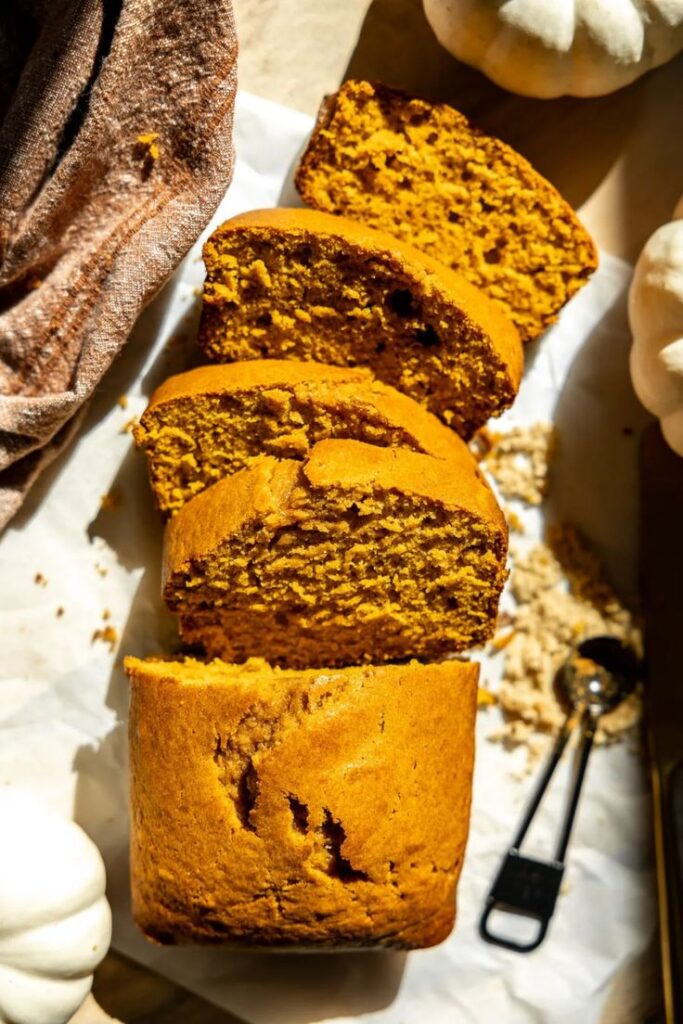 Sliced pumpkin bread on parchment with scattered brown sugar and measuring spoons, surrounded by small pumpkins.