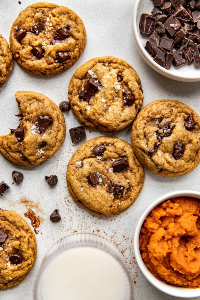 Freshly baked chocolate chip cookies with sea salt, surrounded by chocolate and pumpkin puree on a light surface.
