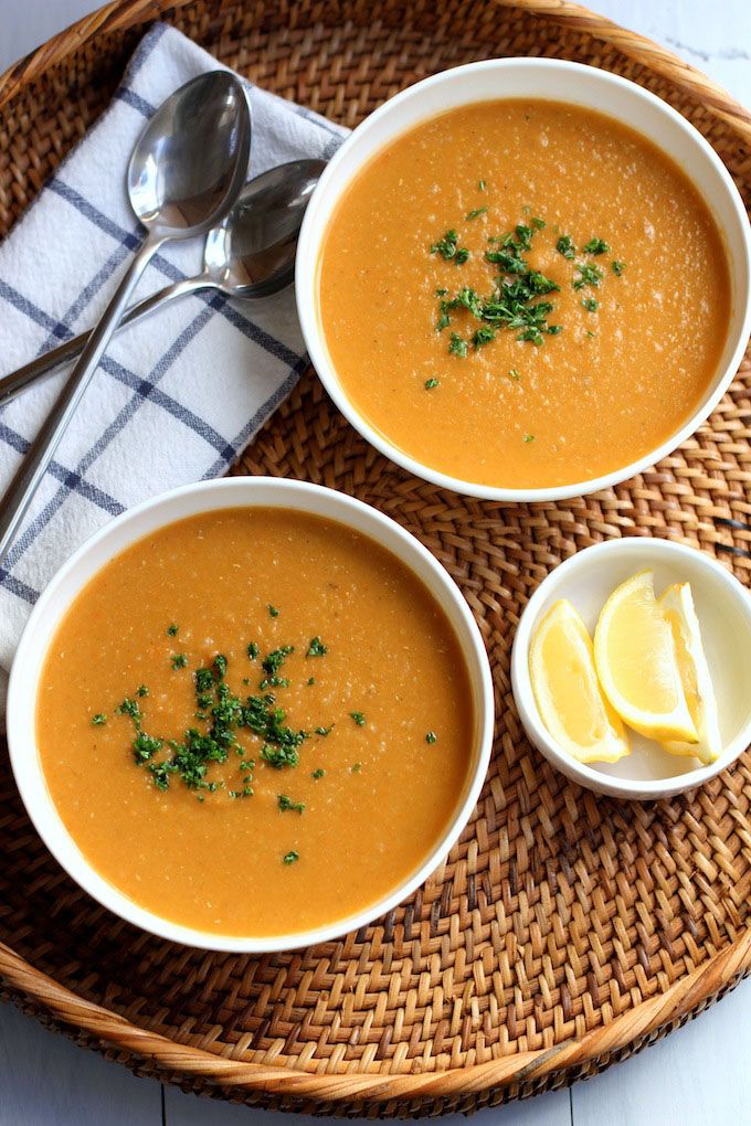 Bowl of lentil soup garnished with herbs, served with lemon wedges and spoons on a wicker tray.