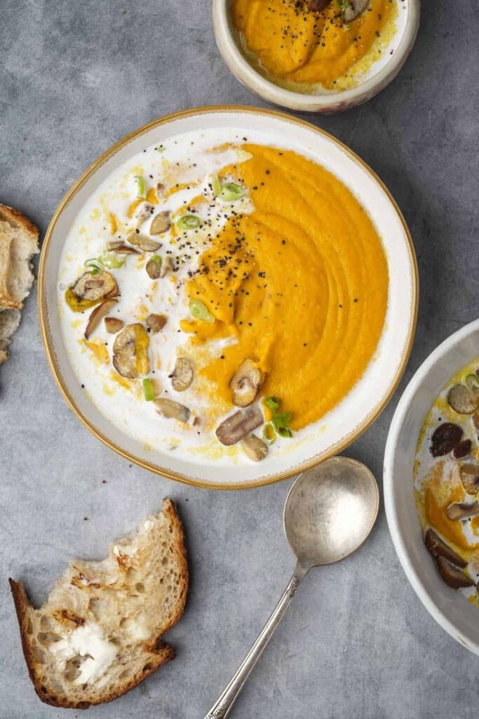 Creamy carrot soup with mushrooms, garnished with herbs, served in a bowl with a side of crusty bread and a spoon.