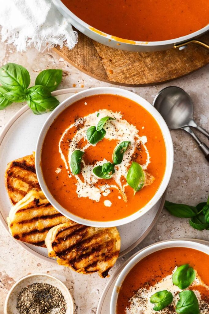 Creamy tomato soup garnished with basil leaves, served with grilled cheese sandwiches on a stone countertop.