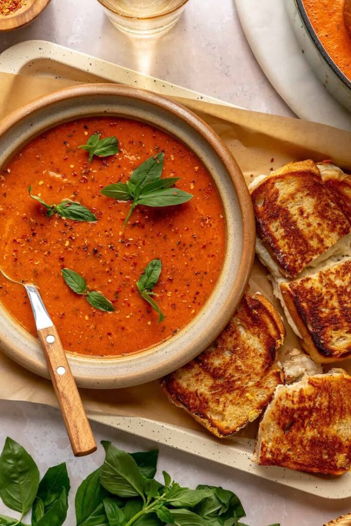 Creamy tomato soup with fresh basil, paired with crispy grilled cheese sandwiches on a tray.