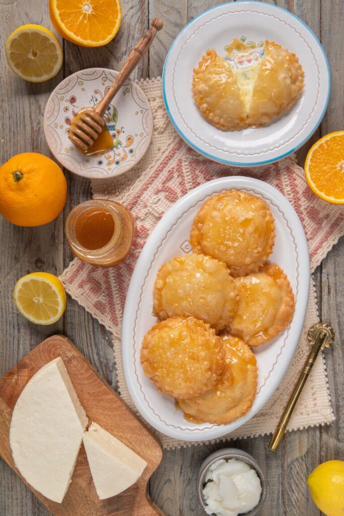 Golden honey pastries on plates with cheese, honey, and fresh oranges and lemons on a rustic wooden table.