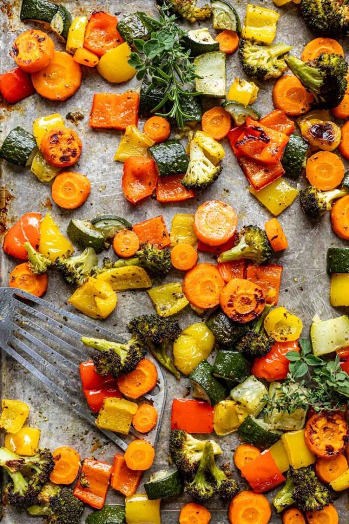 Roasted carrots, bell peppers, zucchini, and broccoli on a baking sheet with herbs and a spatula.