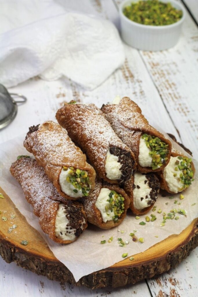 Delicious Sicilian cannoli filled with ricotta, garnished with pistachios and chocolate, served on wooden platter.
