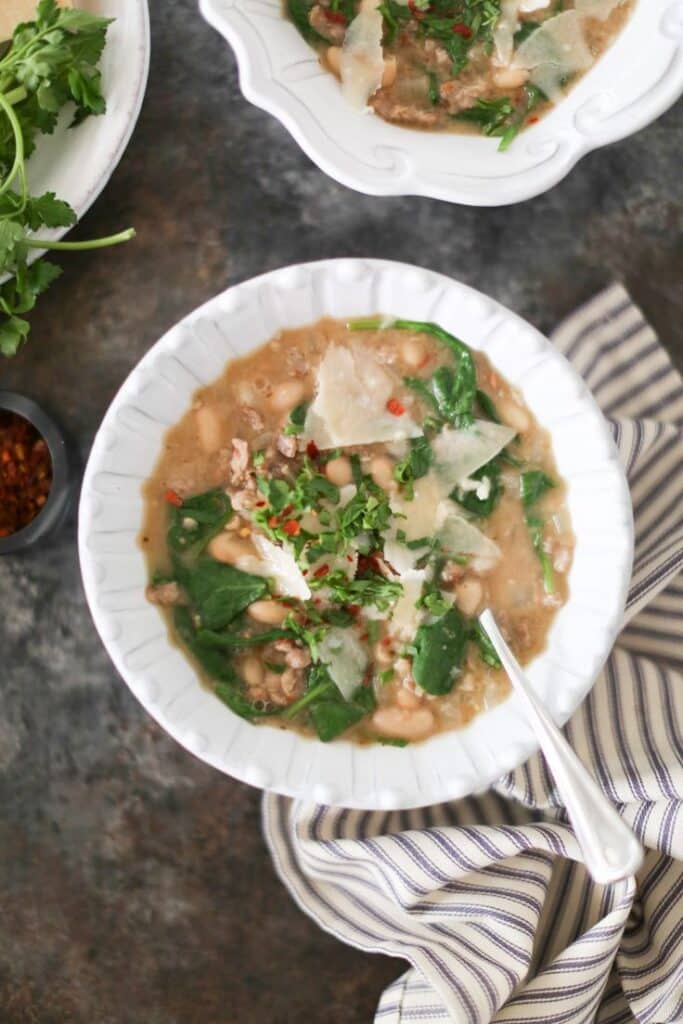 Hearty white bean and spinach soup garnished with herbs and parmesan in a white bowl with spoon on a rustic table.