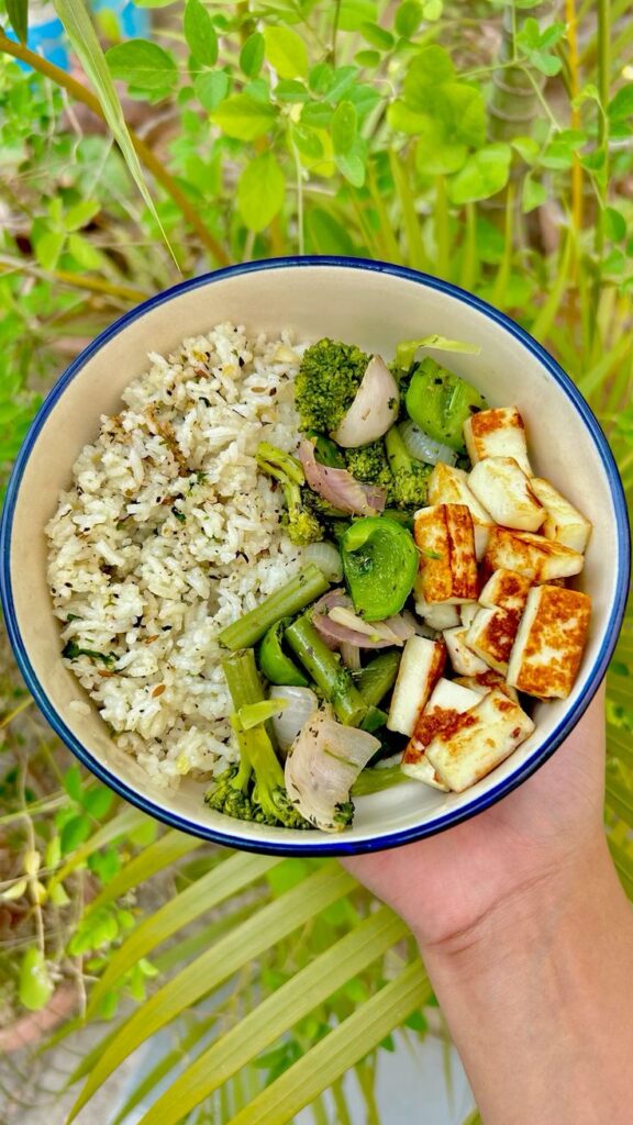 Healthy bowl with rice, grilled paneer, broccoli, and bell peppers, held against a lush green background.