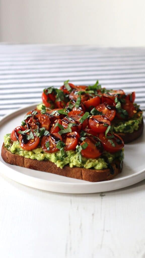 Avocado toast topped with cherry tomatoes, fresh basil, and balsamic glaze on a white plate.