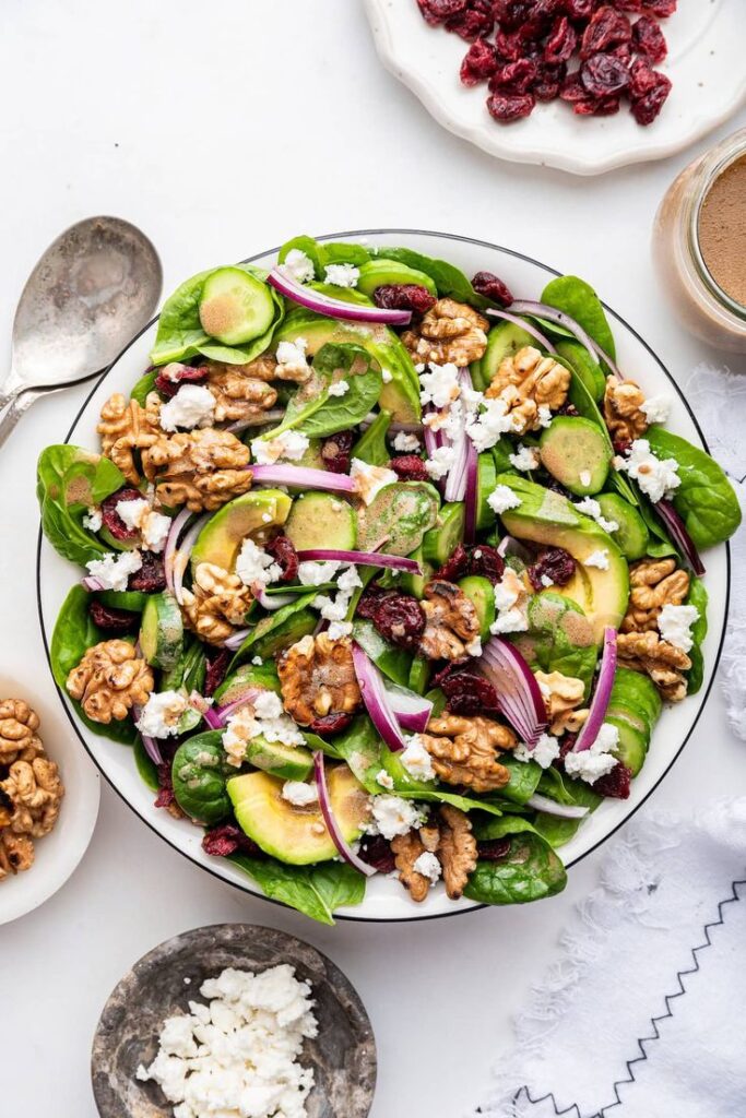 Fresh spinach salad with walnuts, feta, cranberries, cucumber, and vinaigrette in a bowl, on a white background.