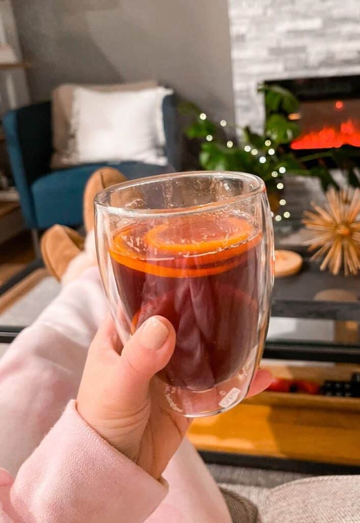 Cozy by the fire with mulled wine in hand, orange slice garnish, and relaxing winter vibes in the living room.