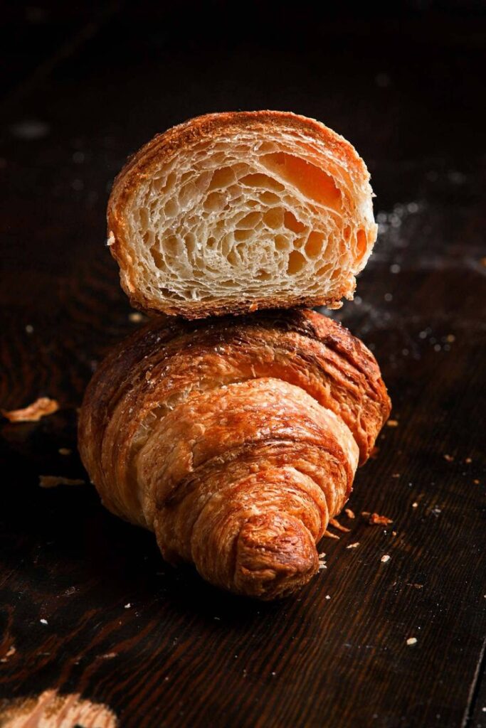 Golden, flaky croissant cut open, revealing airy layers on a dark wooden surface.