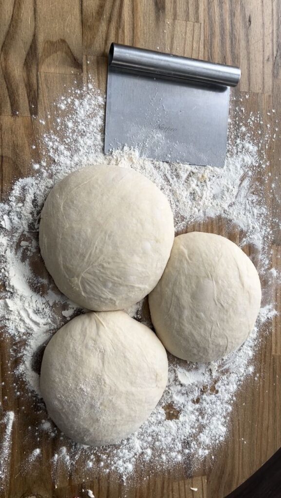 The Perfect Pizza Dough Recipe - Master the Art of Homemade Pizza with Fun, Simple Steps | Fresh pizza dough balls on a floured wooden surface with a metal dough scraper nearby.