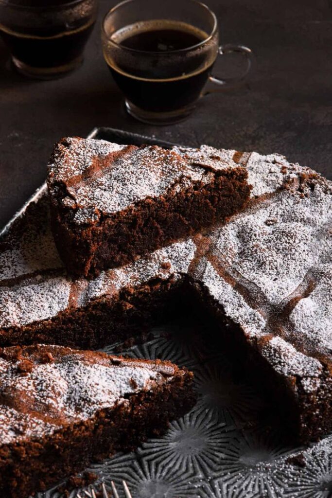 Rich chocolate cake slices dusted with powdered sugar, served with coffee on a dark plate.