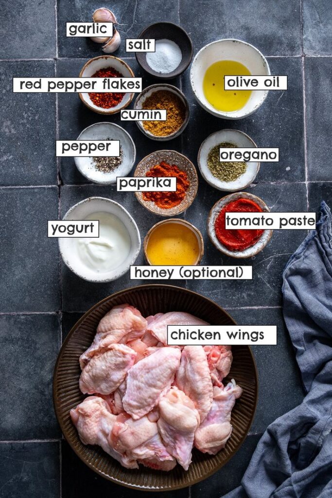 Ingredients for marinated chicken wings: garlic, olive oil, spices, yogurt, tomato paste, and honey on a slate surface.