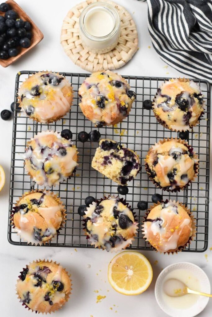Blueberry lemon muffins with glaze on a cooling rack, surrounded by fresh blueberries and lemon slices.